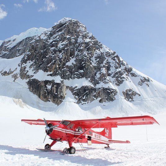 Red airplane on skis parked on a snowy plain in front of a snow-covered mountain.