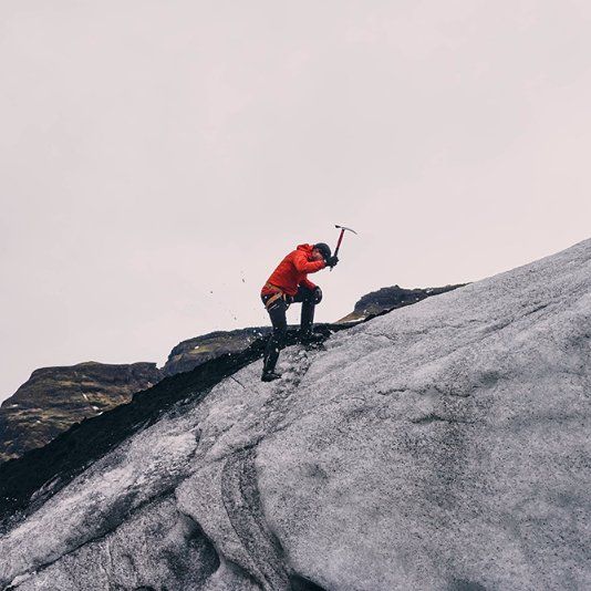 Person in red jacket ice climbing with an ice axe on a glacier, overcast sky.