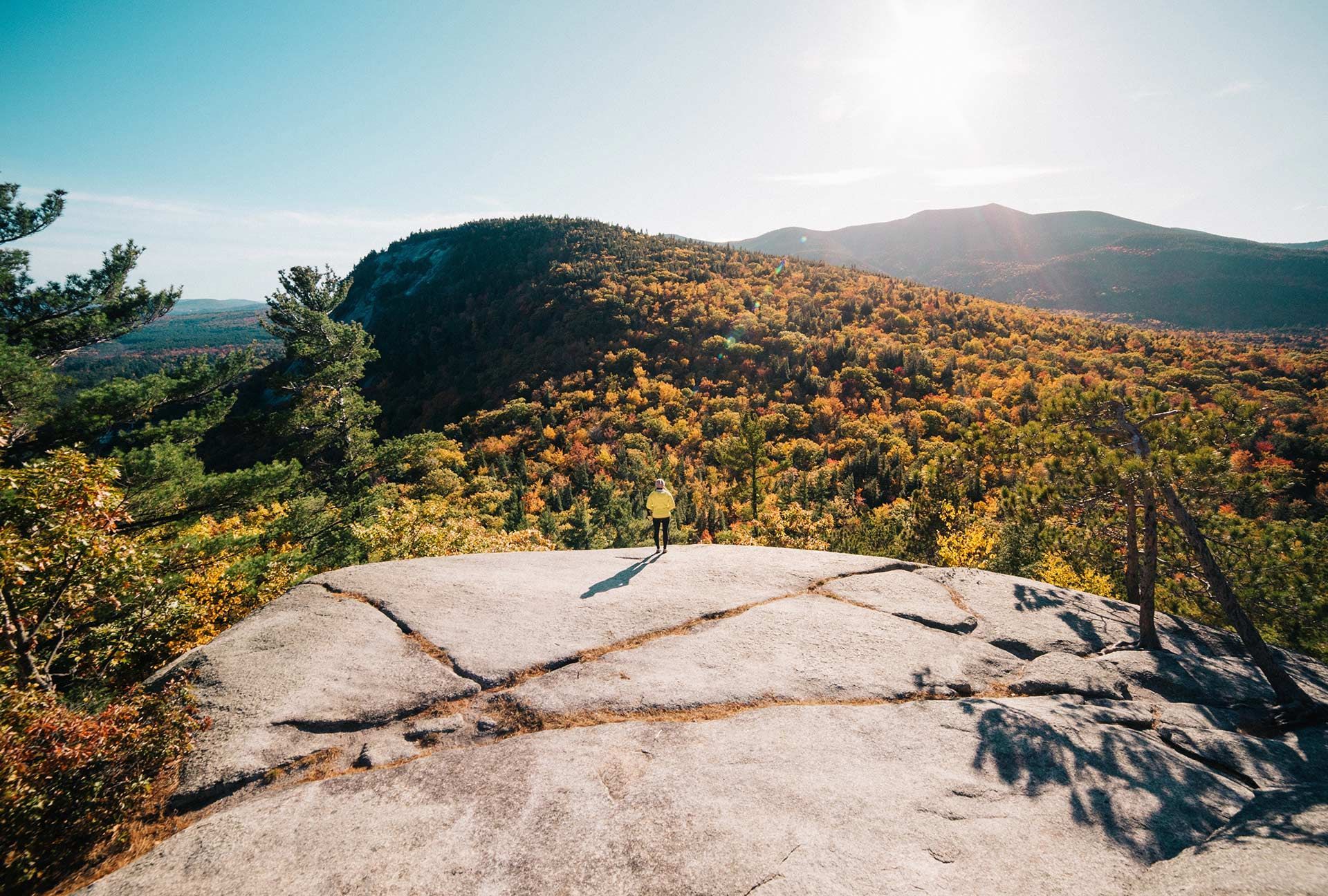 Person stands on a rocky summit, looking at autumn foliage covering mountain slopes under a sunny sky.