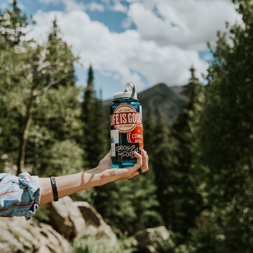 Hand holding a blue water bottle with stickers against a mountain landscape.