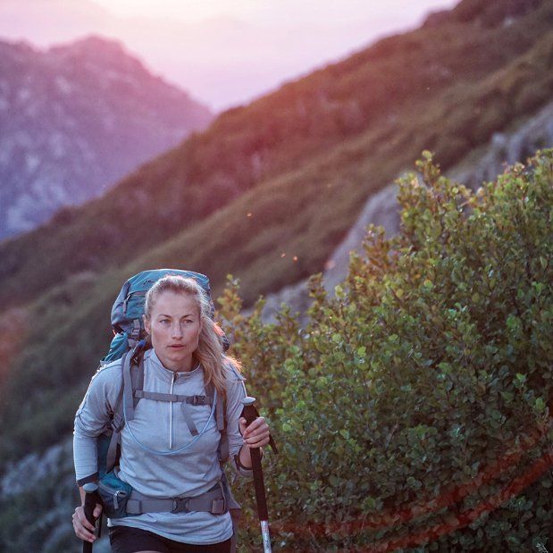 Woman hikes uphill with backpack and trekking poles in a mountain landscape at sunset.