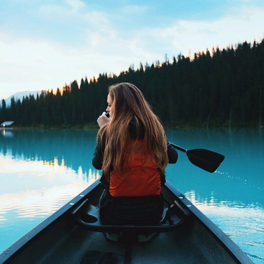 Woman canoeing on turquoise lake, surrounded by forest, sunset in background.