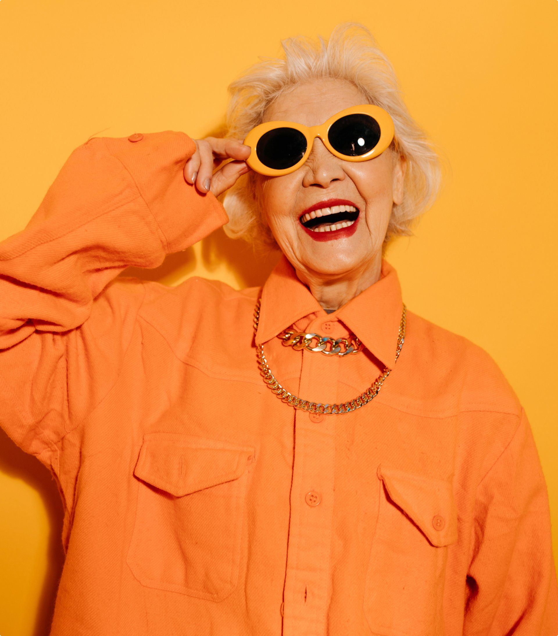 An elderly woman wearing an orange shirt and yellow sunglasses is laughing.