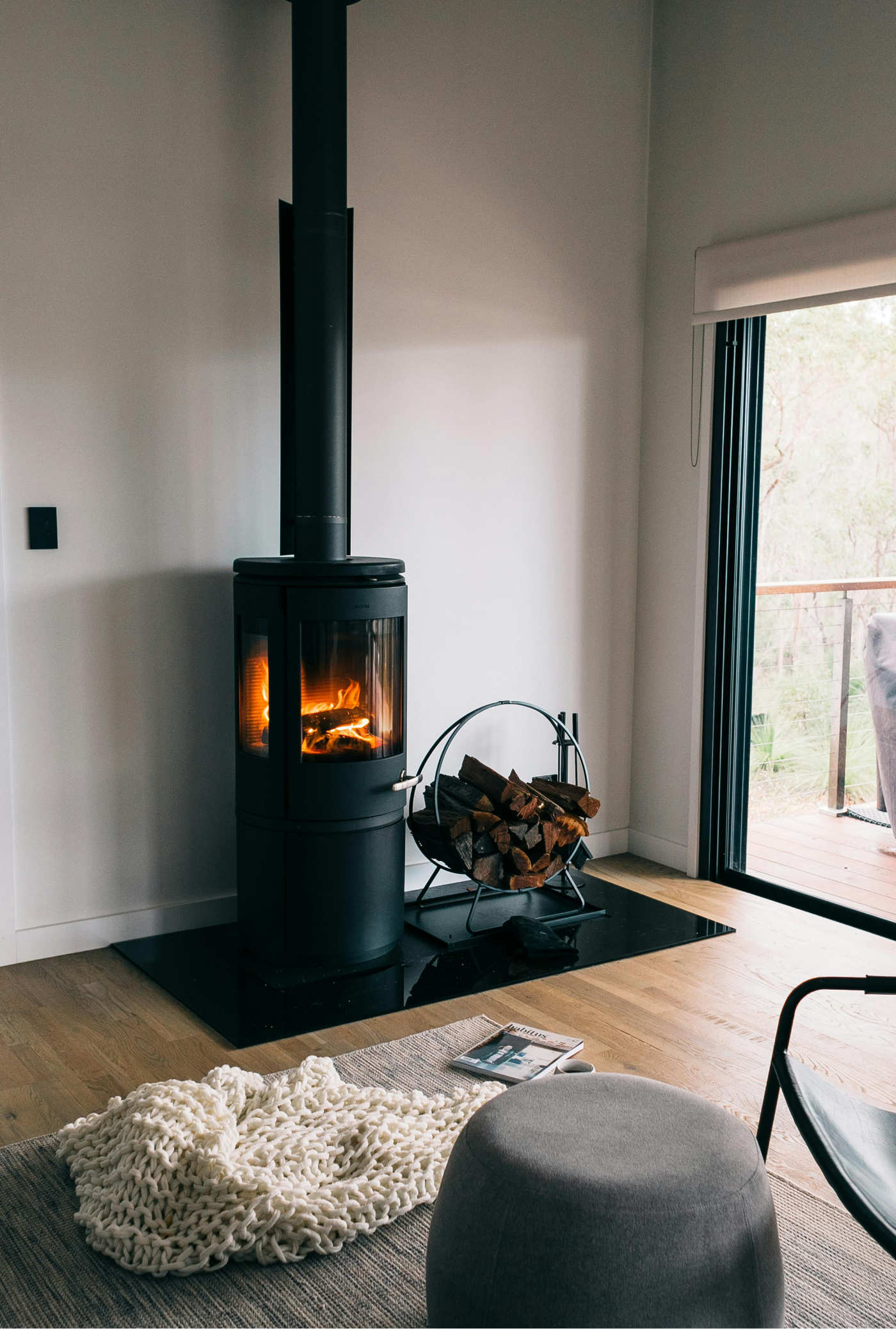 Black stove with lit fire, logs, and a cozy interior. Window view of greenery.