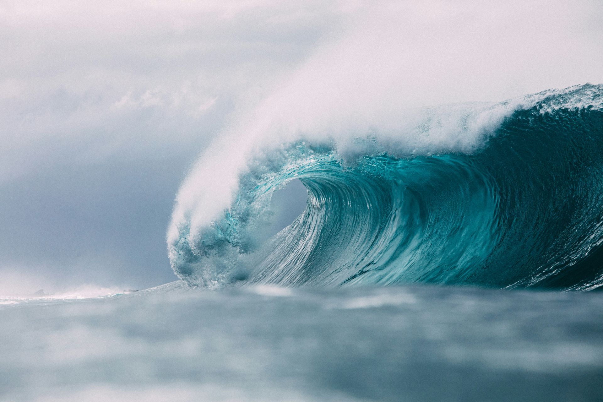 Large, turquoise ocean wave cresting, white foam, under a cloudy sky.