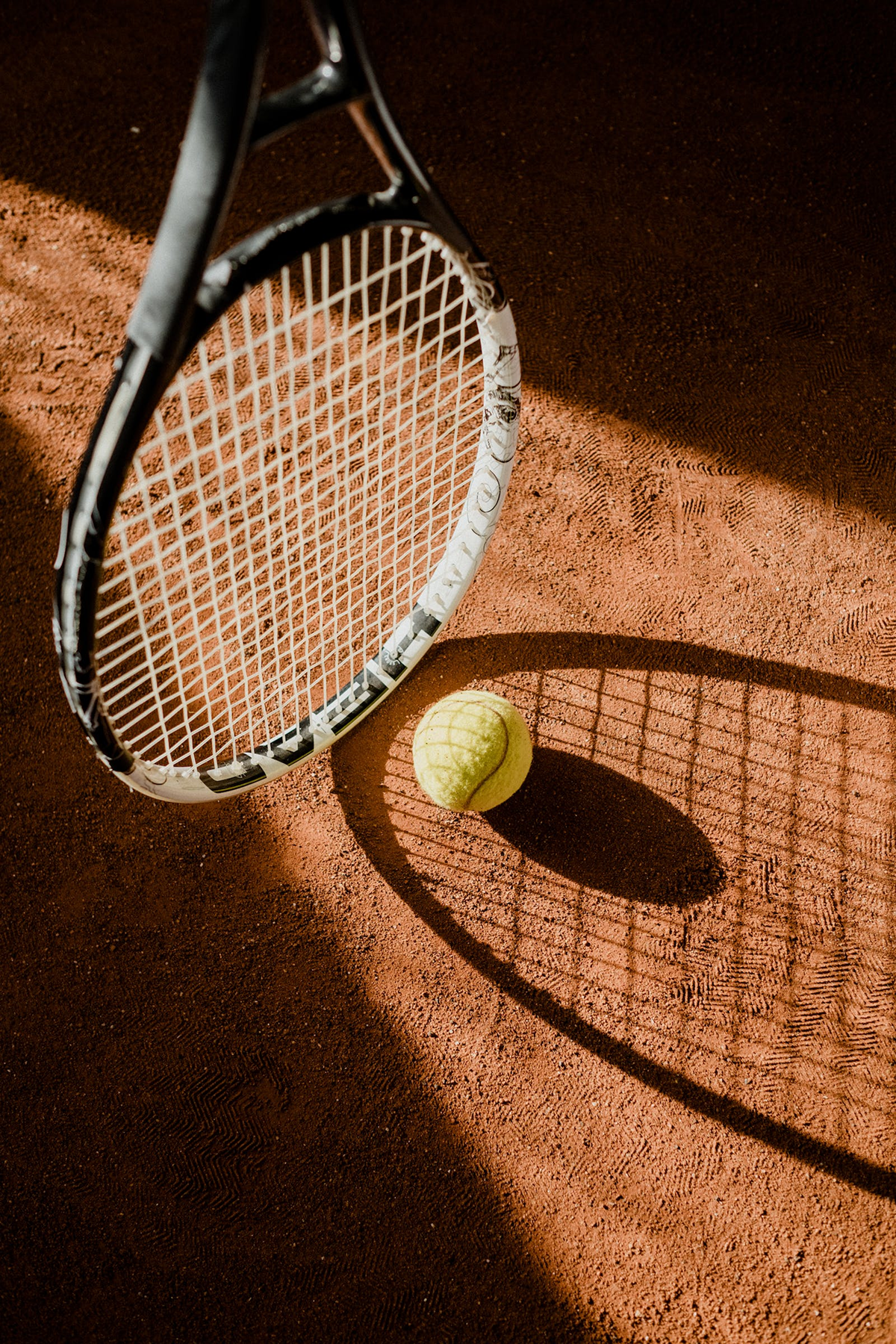 Tennis racket and ball on a clay court, casting long shadow.
