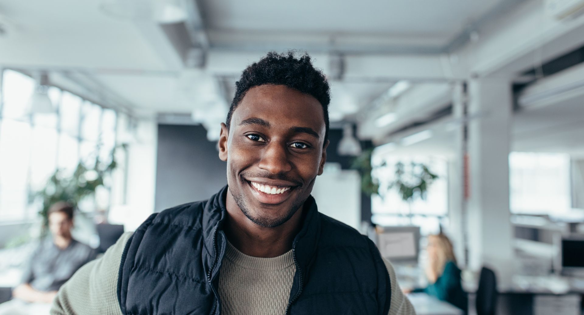 Smiling man in a vest and sweater in a modern office with other people in the background.