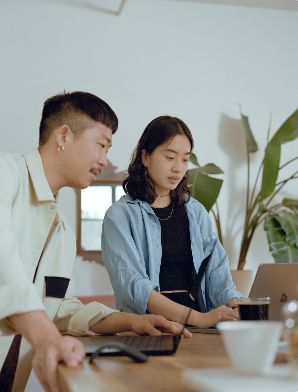 Two people working on laptops at a table, looking at the screen. One wears a light jacket, the other a blue shirt.