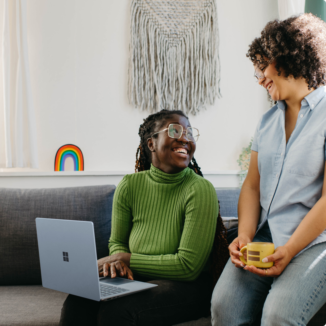Two people on a couch smiling, one using a laptop, the other holding a yellow mug. A rainbow decorates the wall.