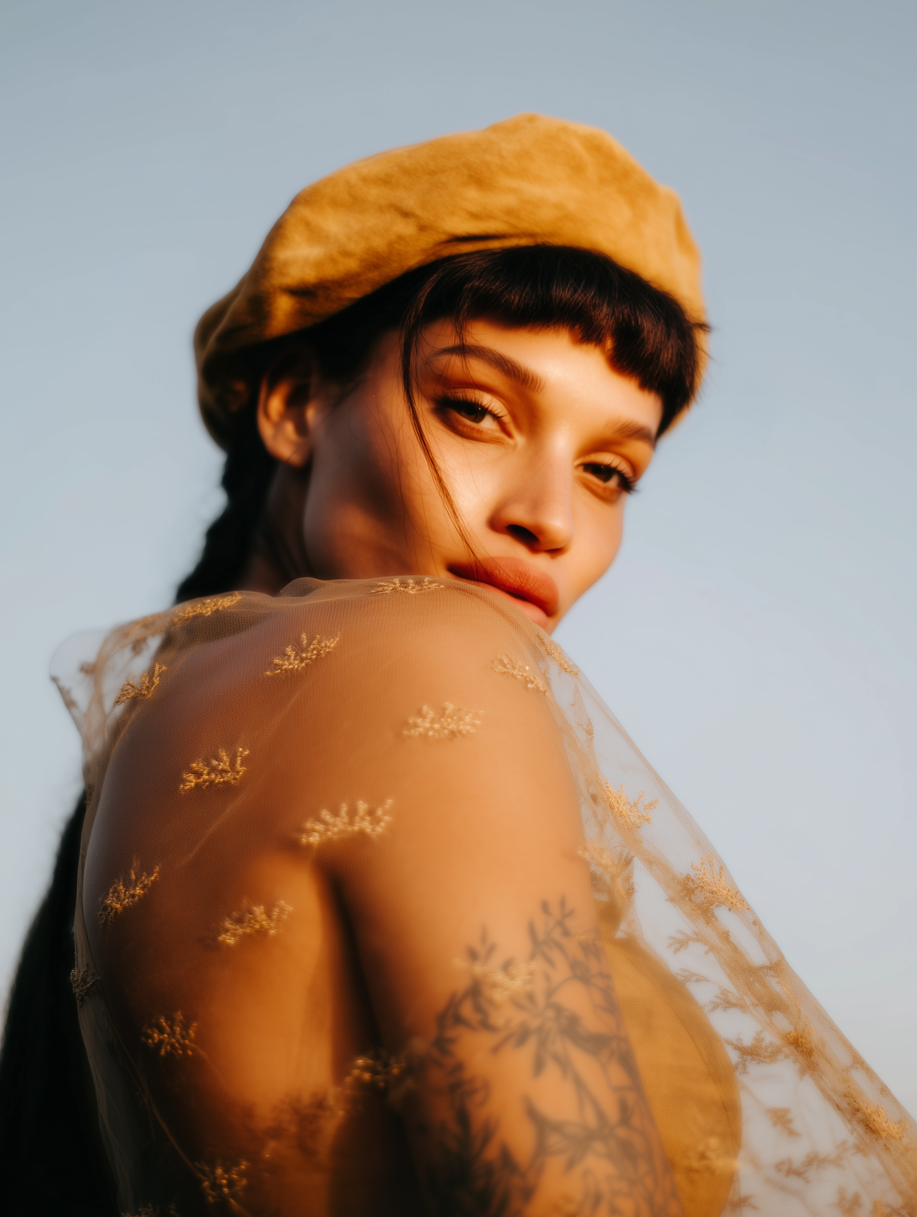 Mulher com boina amarela e blusa transparente com detalhes florais, olhando por cima do ombro; tatuagens visíveis.