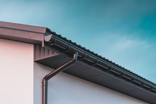 A close-up view of the corner of a roof featuring dark gray metal roofing, dark gutters, and a light-colored wall.