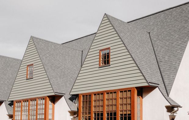 A close-up of a house with grey shingled roofs featuring multiple peaked gables, tall wooden window frames, and light walls.