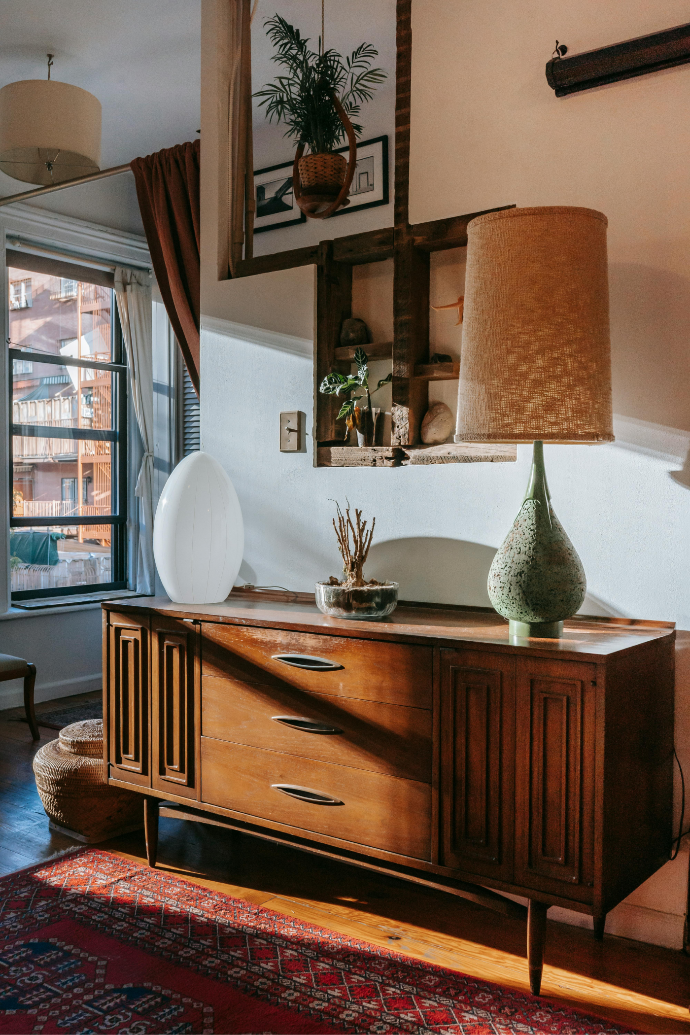 Mid-century modern wooden sideboard with a textured lamp and decor, set against a white wall near a sunlit window.