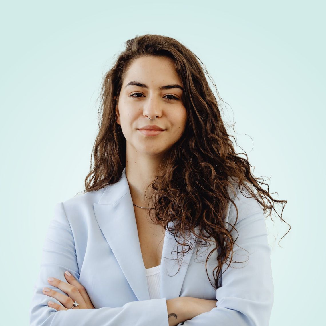 Person with curly dark hair in a light blue blazer, standing with arms crossed against a pale blue background