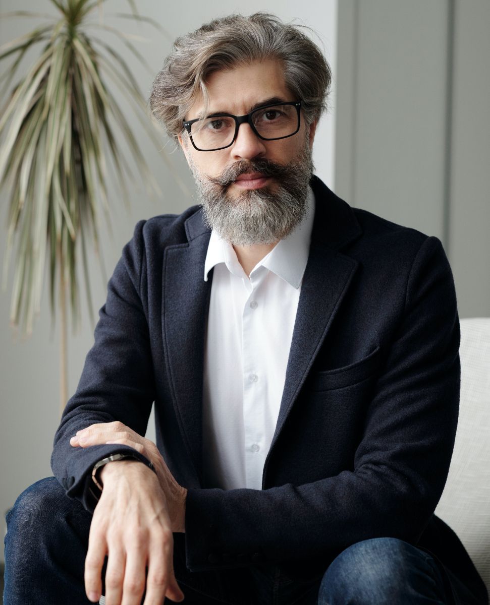 Man with glasses and gray beard in a dark blazer seated on a sofa, looking at the camera.