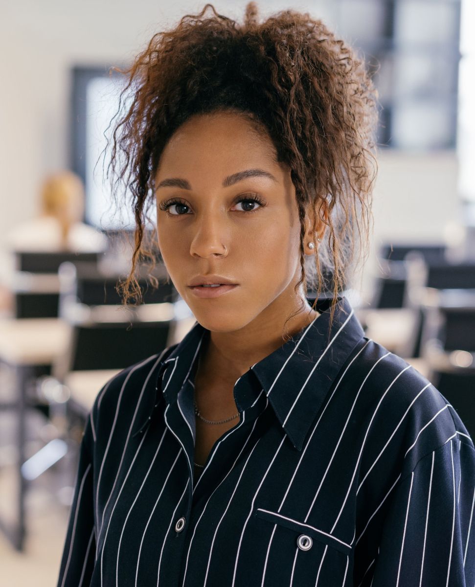 Woman in a striped shirt with curly hair, looking at the camera in a bright office.