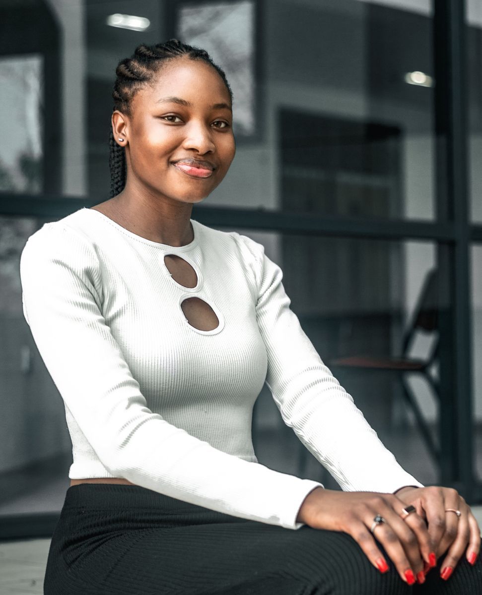 Smiling person in a white top and black pants seated outdoors by a glass building.