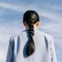 A person with a braided ponytail wears a light blue blazer, viewed from the back against a blue sky with faint clouds.