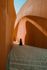 A person in black walks through a path between two towering, textured, orange-brown curved walls under a clear sky.