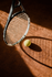 A tennis racket stands over a tennis ball on a clay court, casting a long shadow in the sunlight.