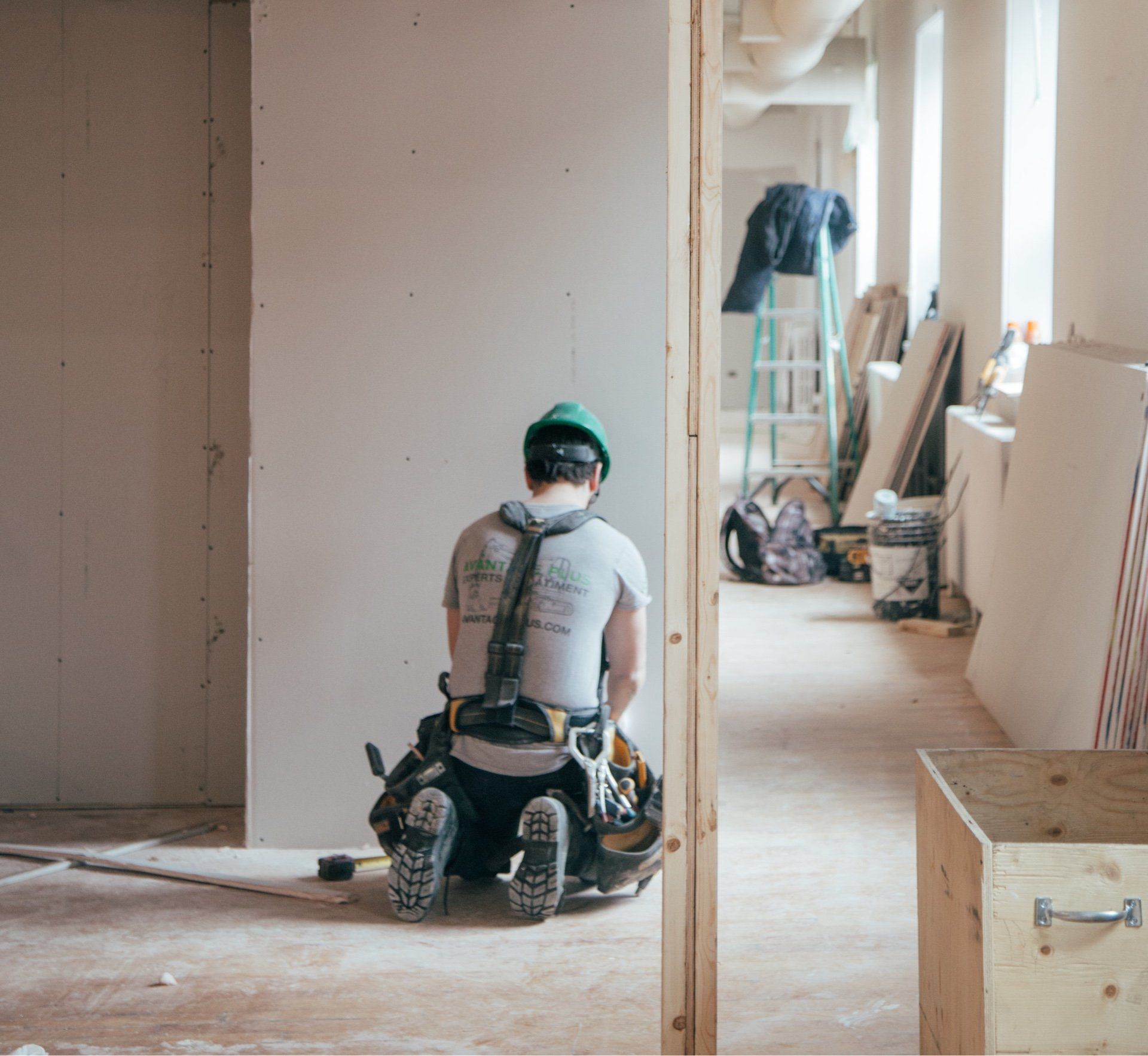 Construction worker kneeling, facing away, in a building under renovation; wearing a green helmet, tool belt.