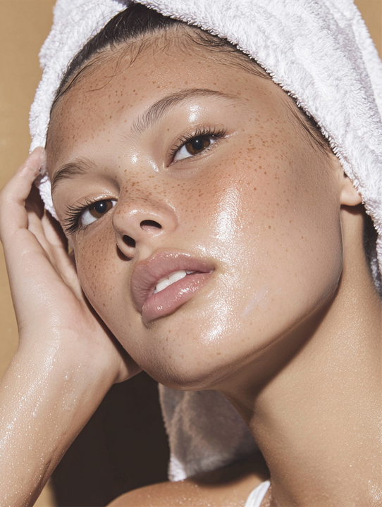 Young woman with a towel on her head, freckles, dewy skin, looking at the camera with a soft expression.