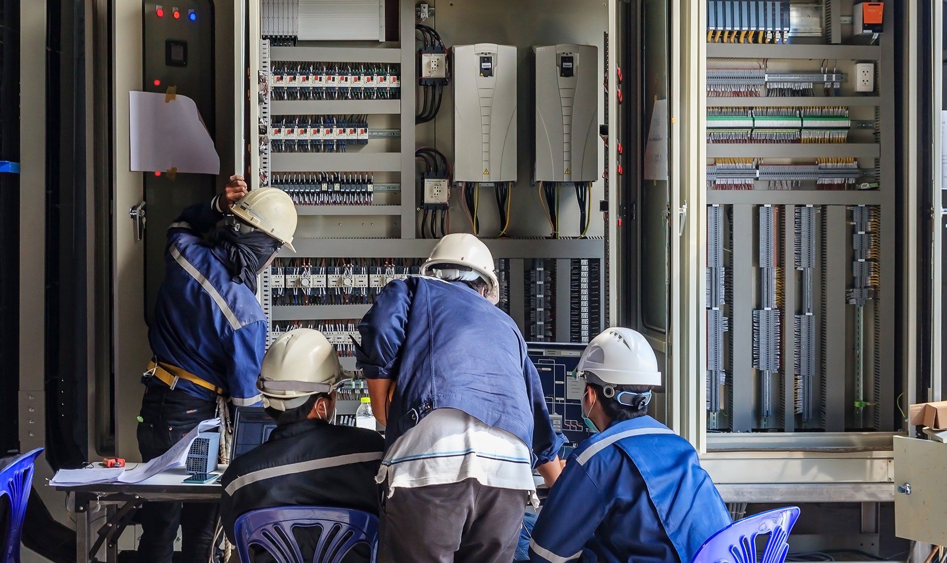 Electrical workers in blue coveralls and hard hats working on an electrical panel.