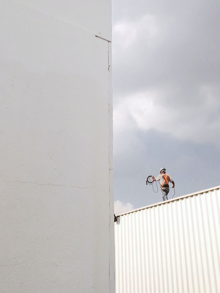 Person standing on a rooftop edge with a camera against a cloudy sky and white building wall