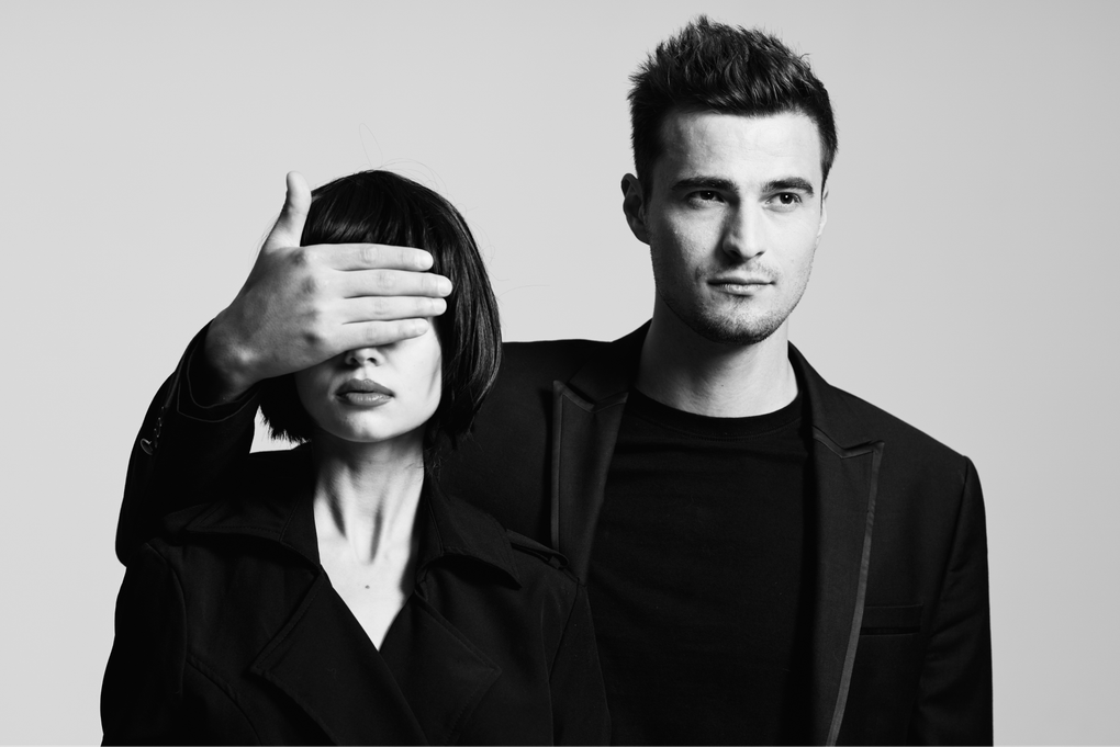 A man and a woman are standing next to each other and the woman is covering the man 's eyes.