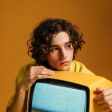 Young person with curly brown hair, resting chin on a small yellow television. Orange background.