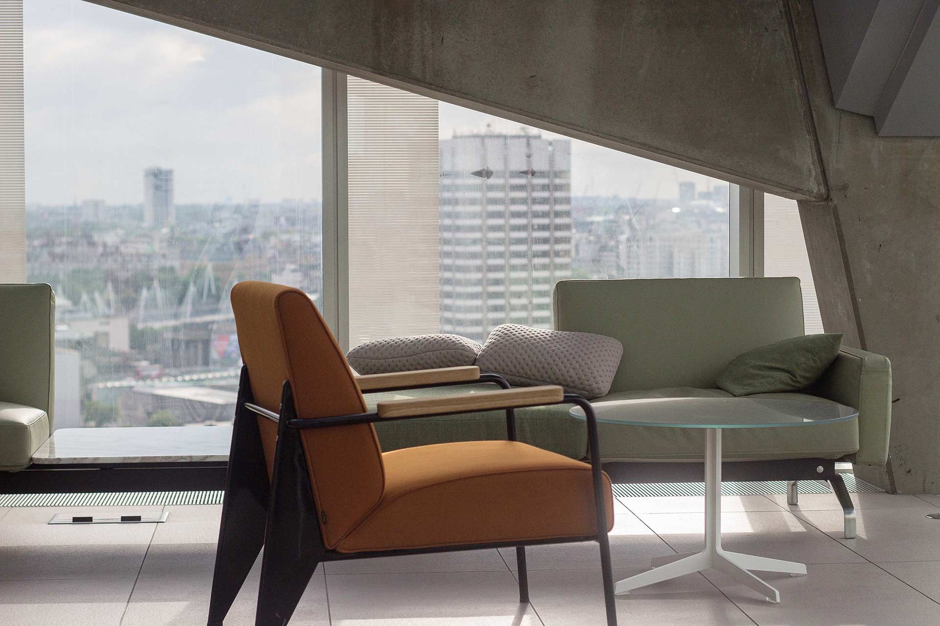 An armchair and a sofa face a large window overlooking a city skyline inside a room with angular concrete walls.