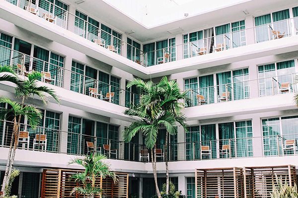 A multi-story hotel courtyard with white balconies, large glass windows, and palm trees reaching toward the open sky.