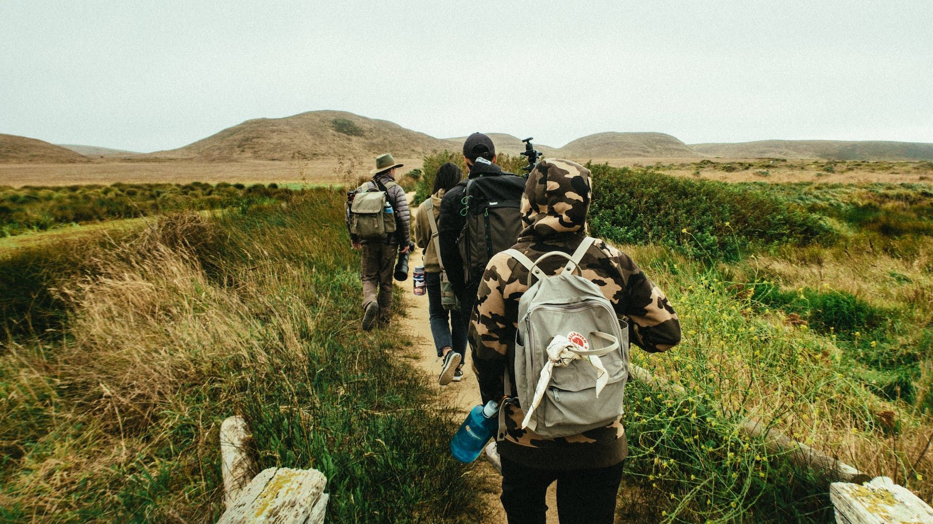 Group of people hiking along a path through tall grass, under a cloudy sky.