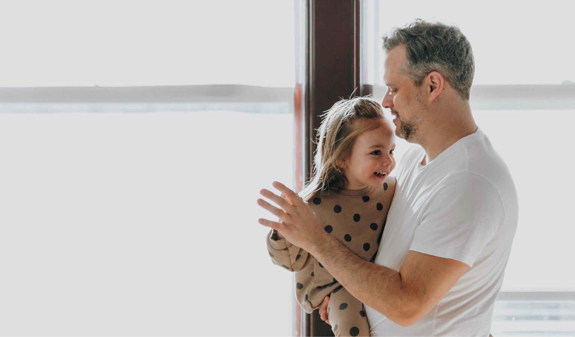 Un hombre sostiene y sonríe a un niño pequeño cerca de una ventana. La luz del sol entra a raudales.