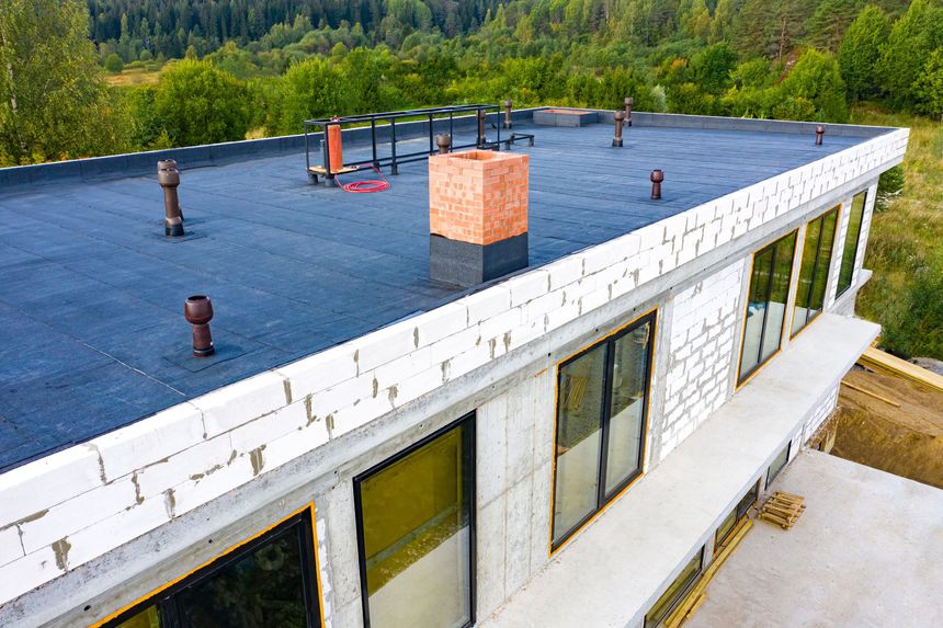 Rooftop of a white building with a dark flat roof, vents, and a brick chimney, surrounded by trees