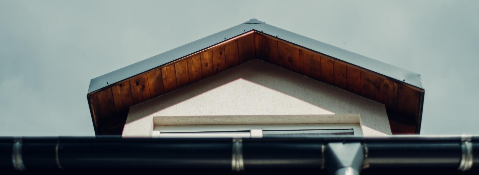 A low-angle shot of a house with a wooden-trimmed roof and a black gutter against a cloudy sky.