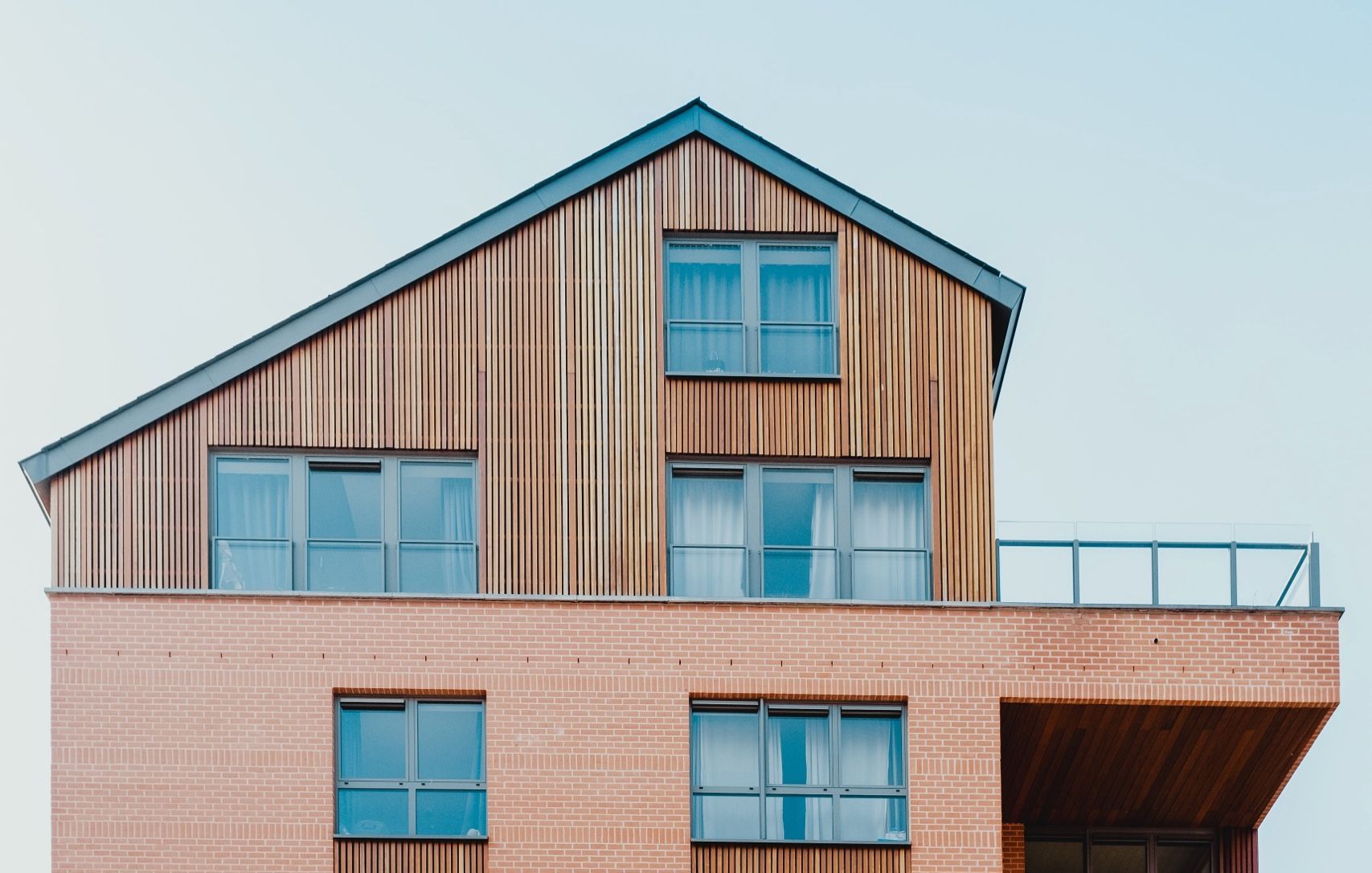 Modern building with brick facade and wooden top story, blue windows, and glass railing.