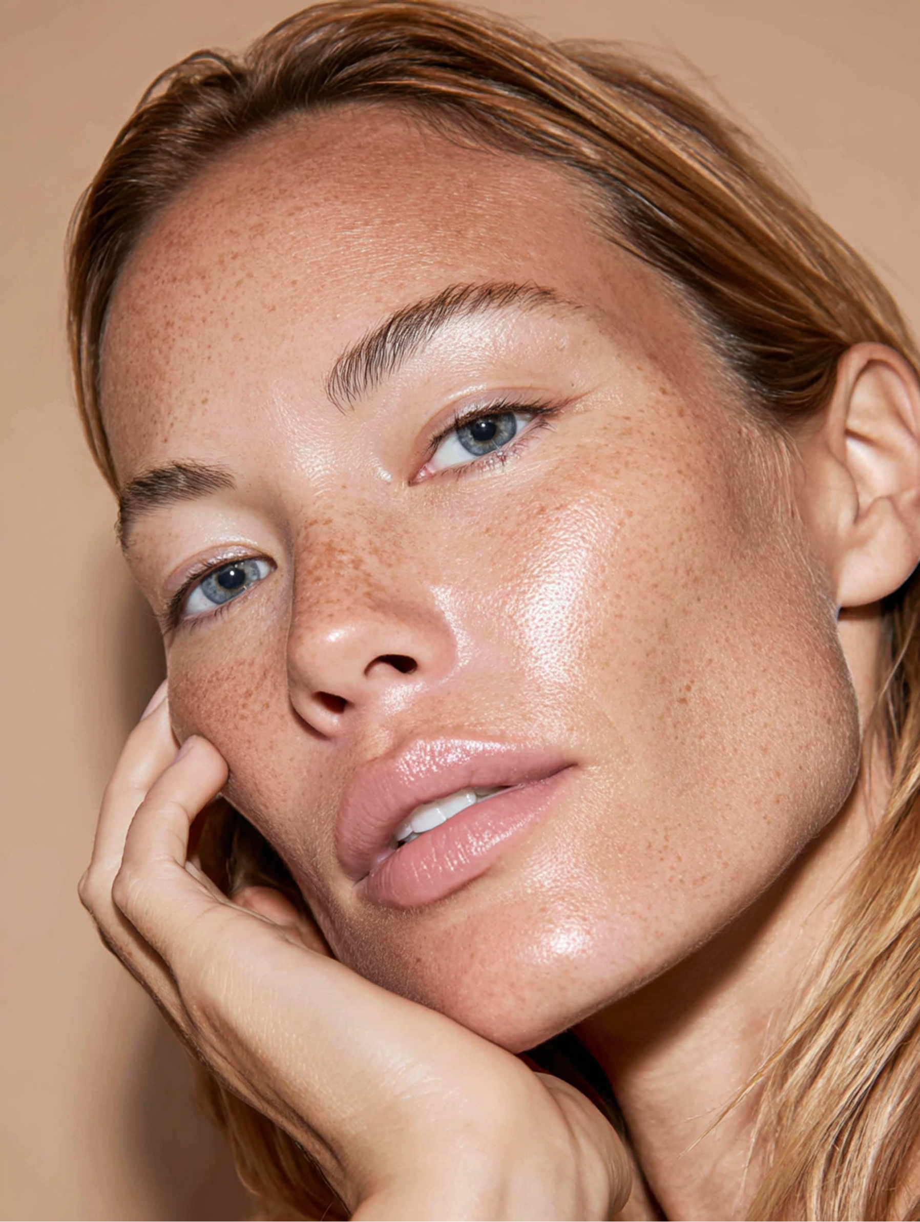 Close-up of a person with freckles, dewy skin, and neutral lip gloss resting their chin on their hand.