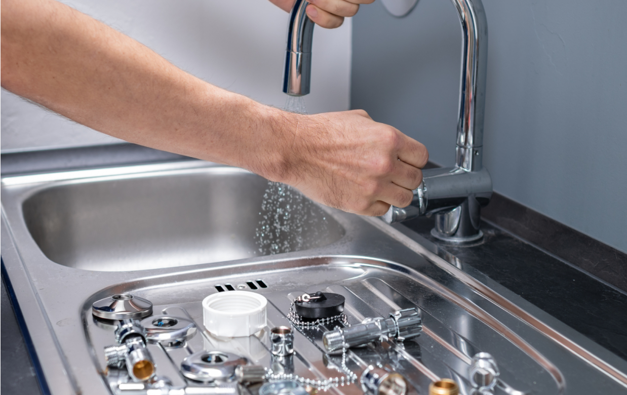 Eine Person testet einen Wasserhahn in einem Edelstahlwaschbecken; daneben liegen Sanitärteile bereit.
