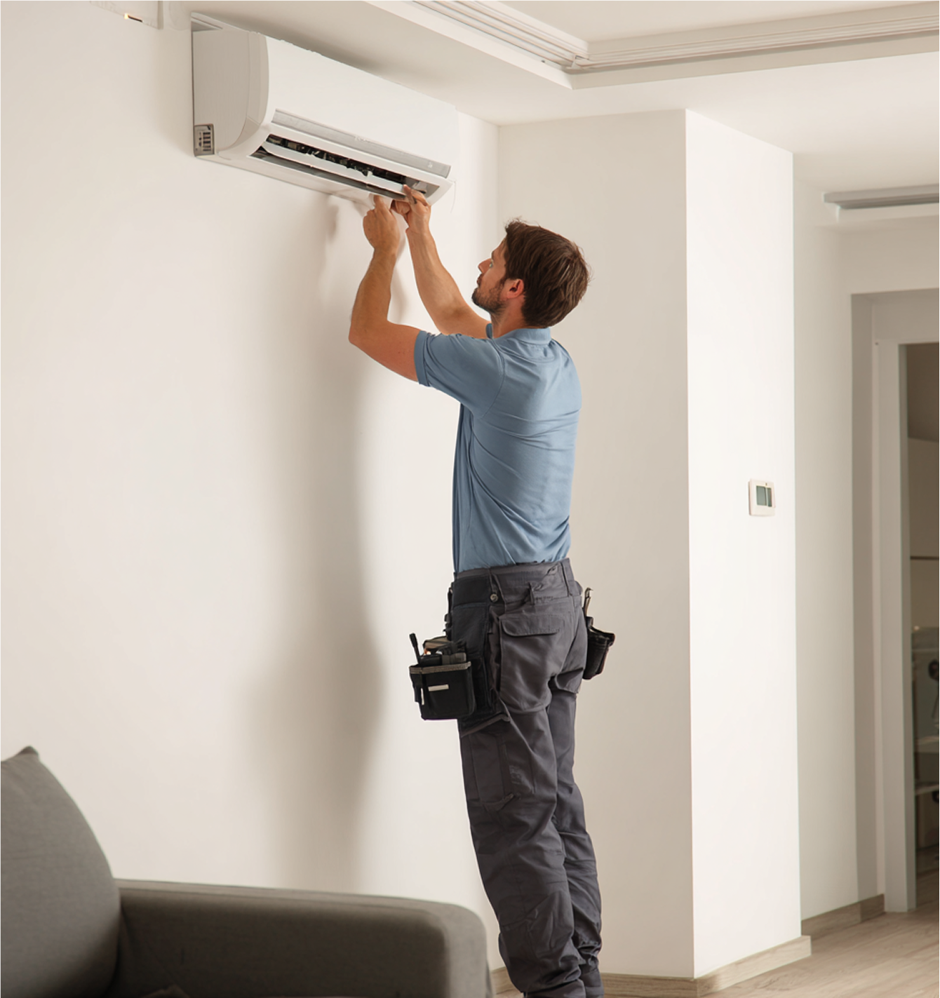 Man servicing air conditioner on a white wall. He is wearing a tool belt.