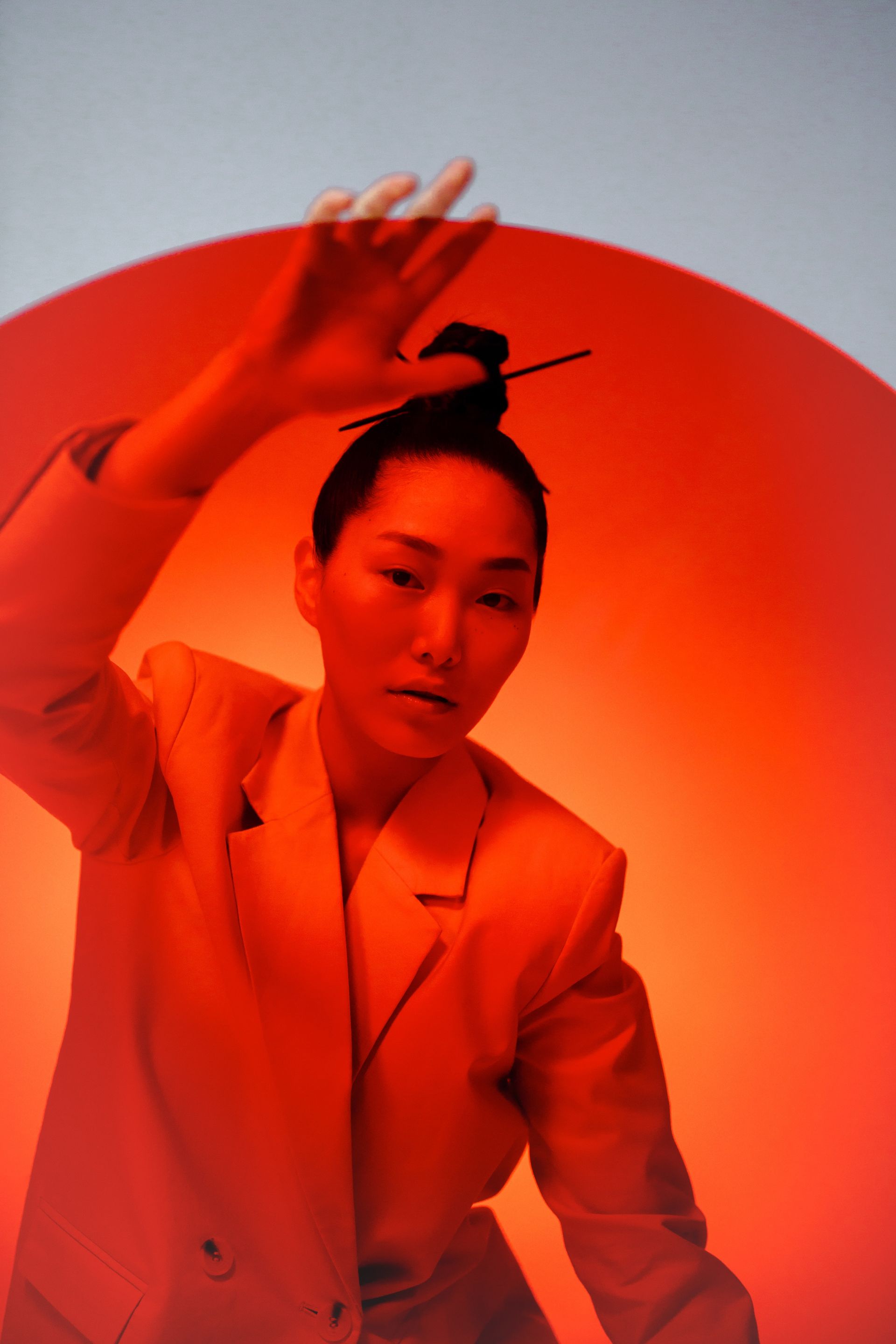 Woman in beige blazer, hand raised, hair bun with pin, against orange and blue gradient background.