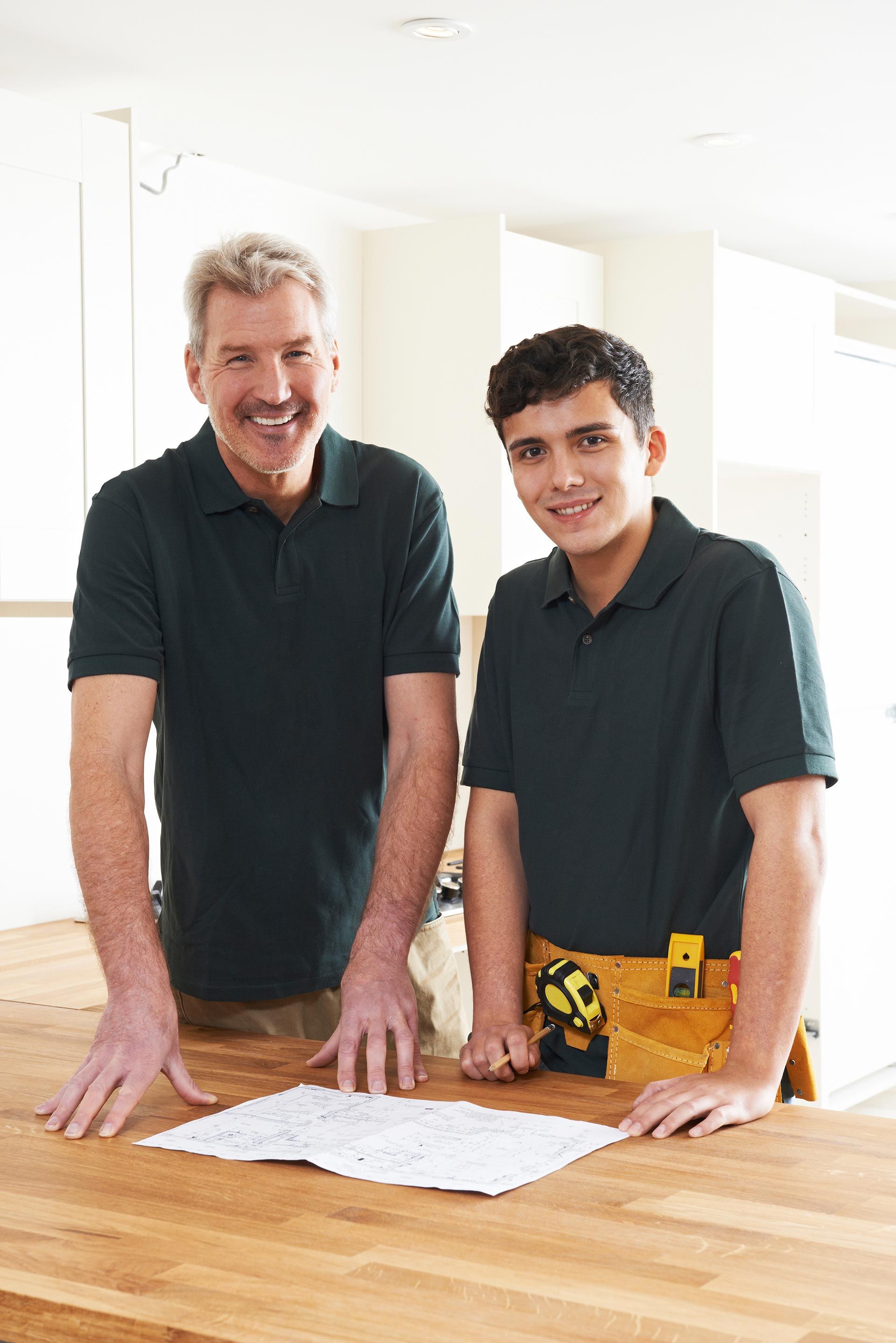 Two people in green shirts reviewing plans on a wooden surface, smiling.