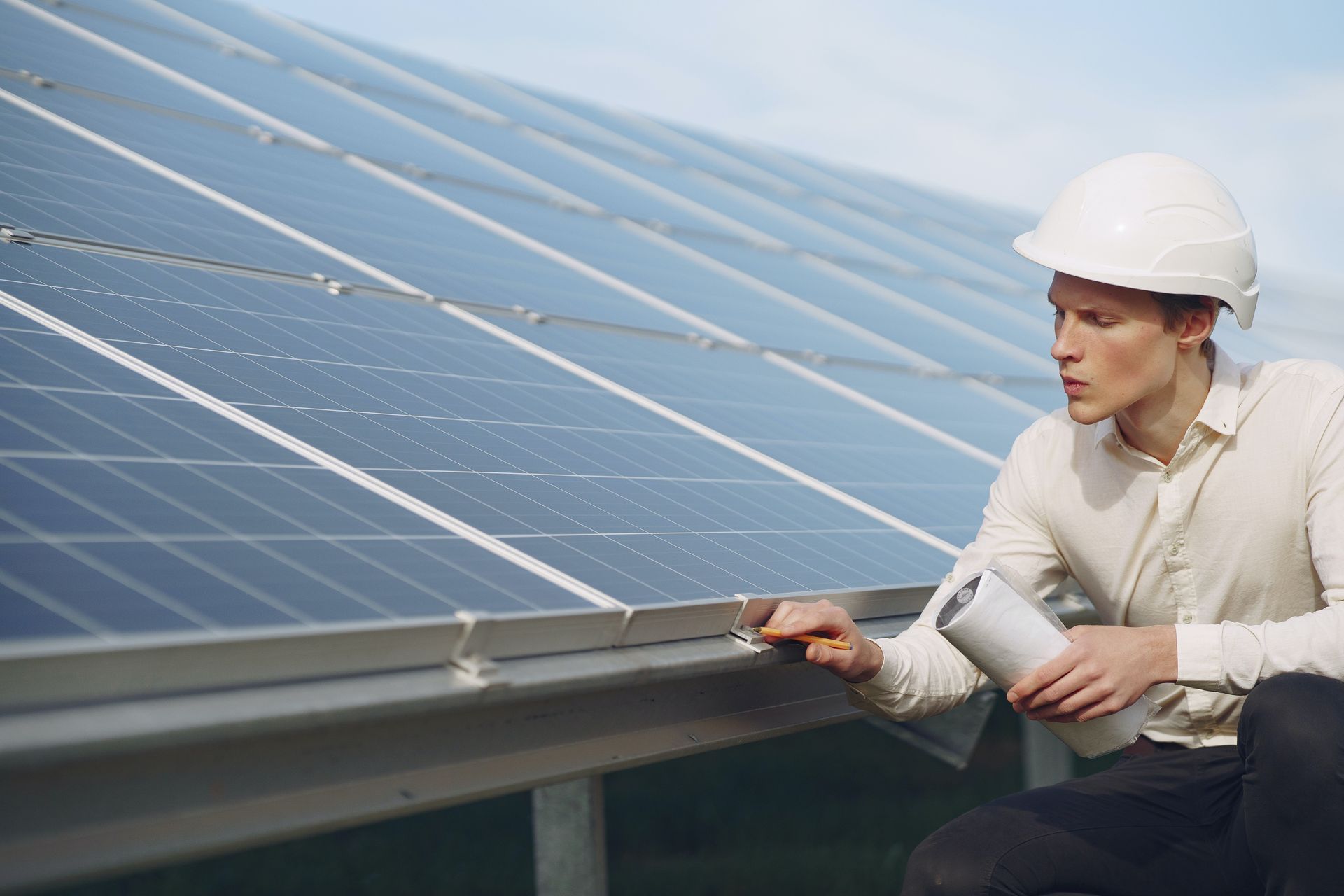 Man in hard hat examines solar panels, holding a cylindrical object outdoors.