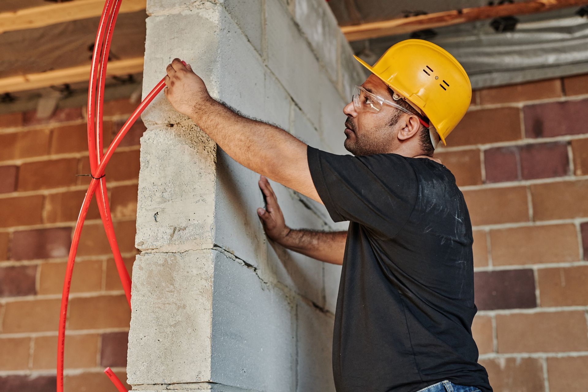 Construction worker in a yellow hard hat, installing red tubing on a cinder block wall.