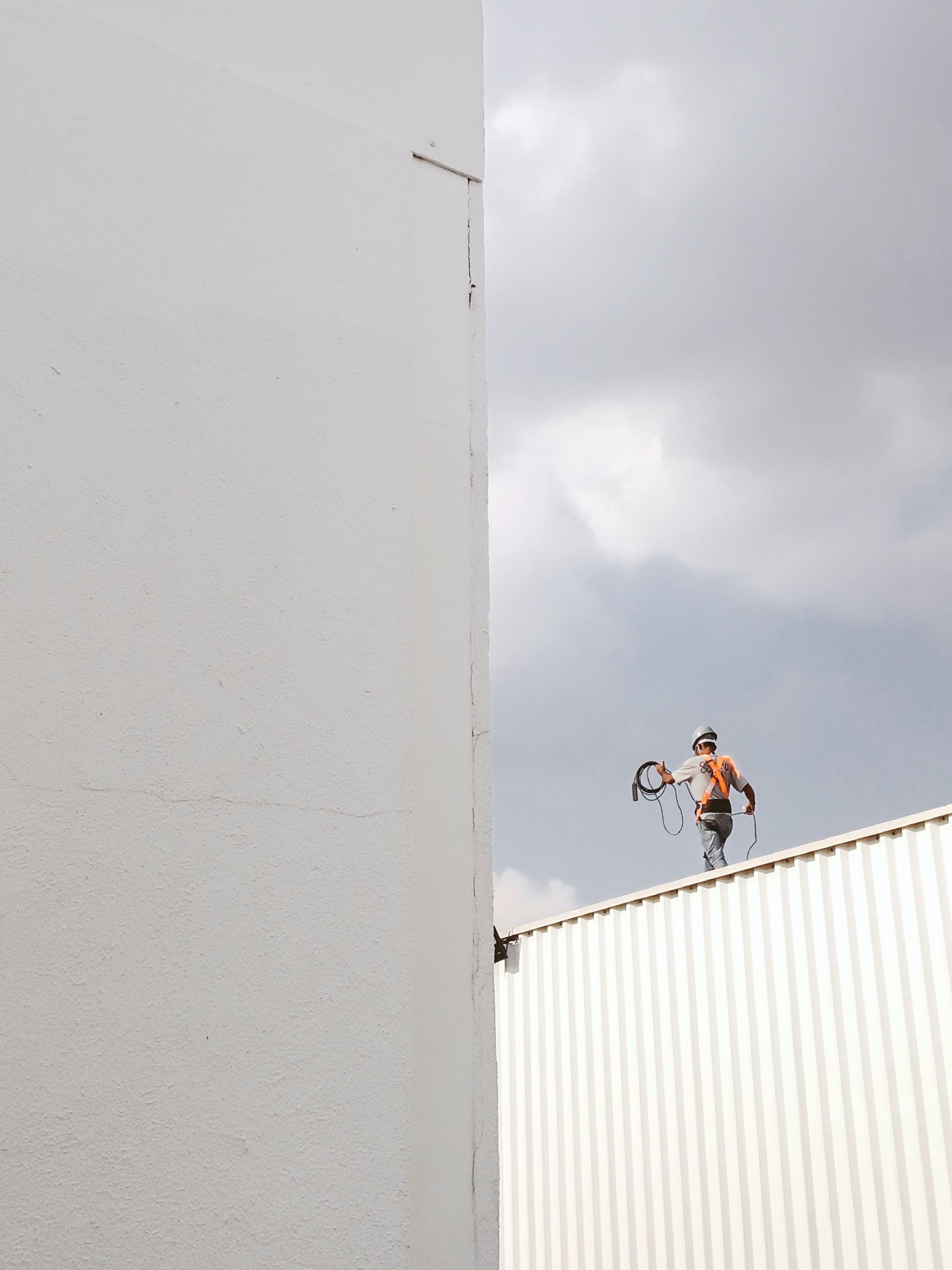 A worker in safety gear stands on a white roof, holding equipment; the sky is cloudy.