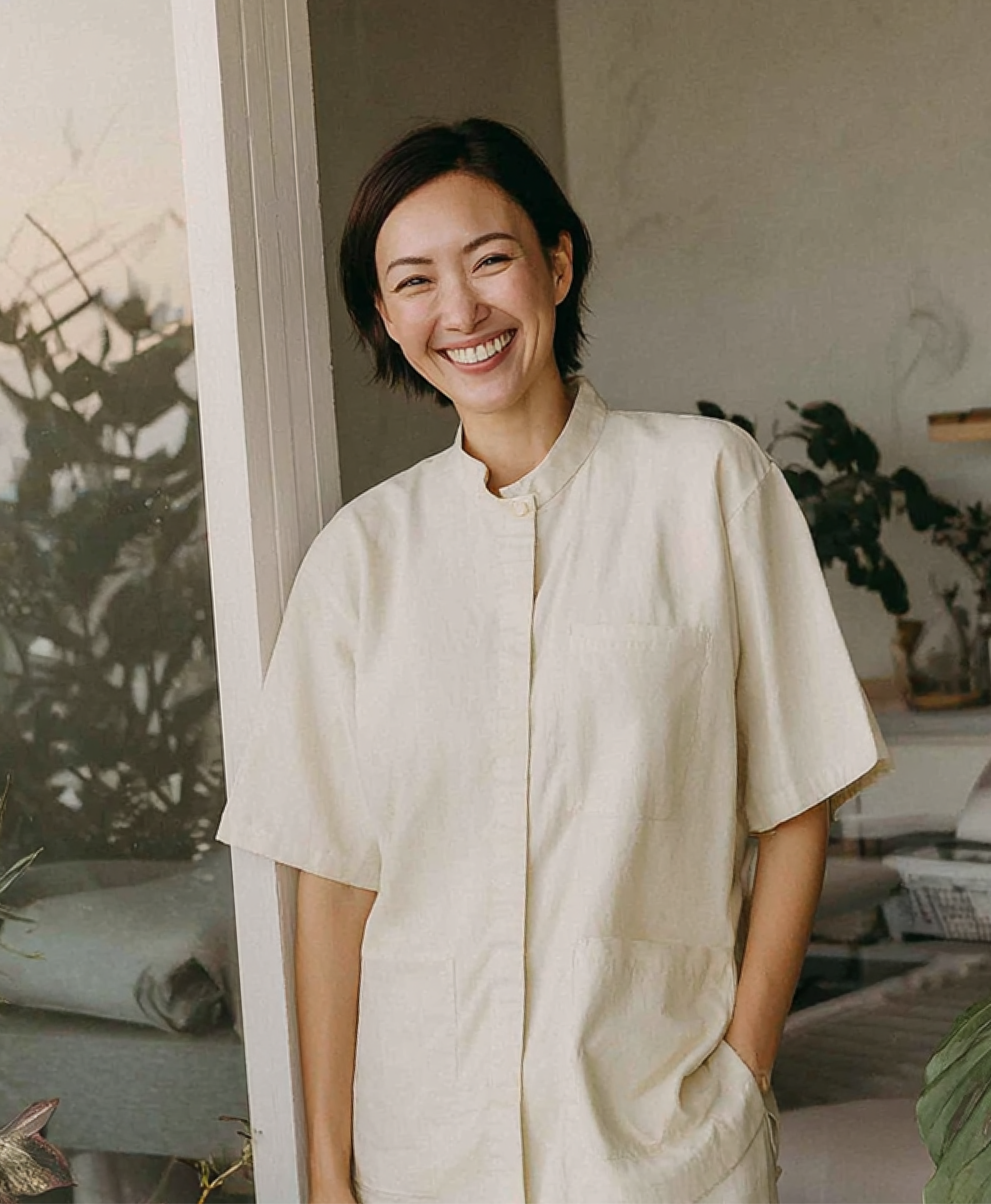 Een glimlachend persoon met kort donker haar draagt een beige overhemd met korte mouwen in een lichte, met planten gevulde kamer.