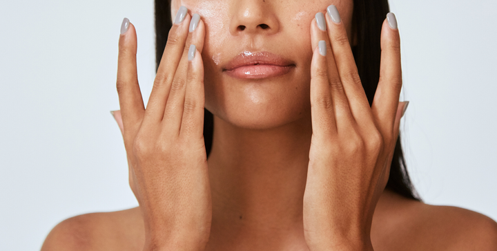 Woman applying skincare product to her face with both hands, light blue backdrop.