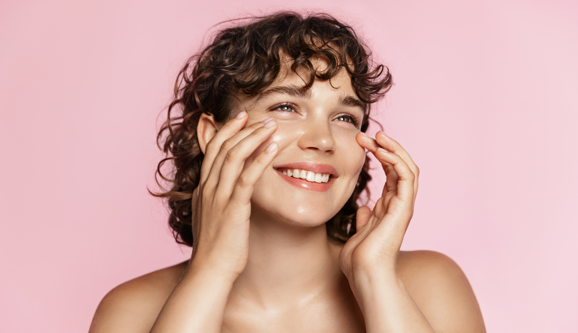Woman with curly hair smiling, applying skincare to her face with both hands, pink background.