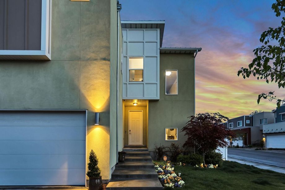 Modern two-story house with green exterior, lit doorway, and garage against a sunset sky.