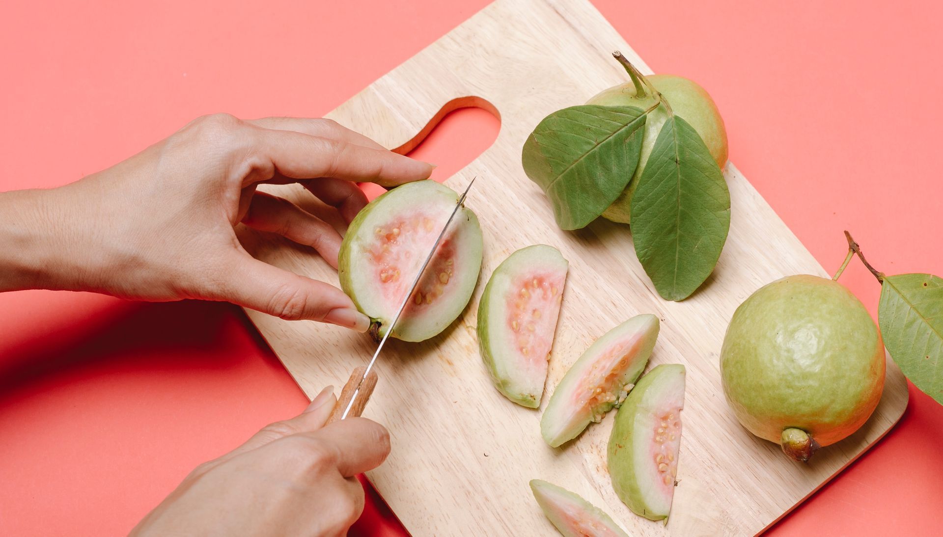 Person slicing guava fruit on a wooden cutting board; pink background.